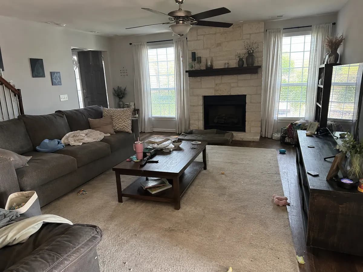 A living room before cleaning, with laundry, papers and clutter across the sofa, coffee table and floor